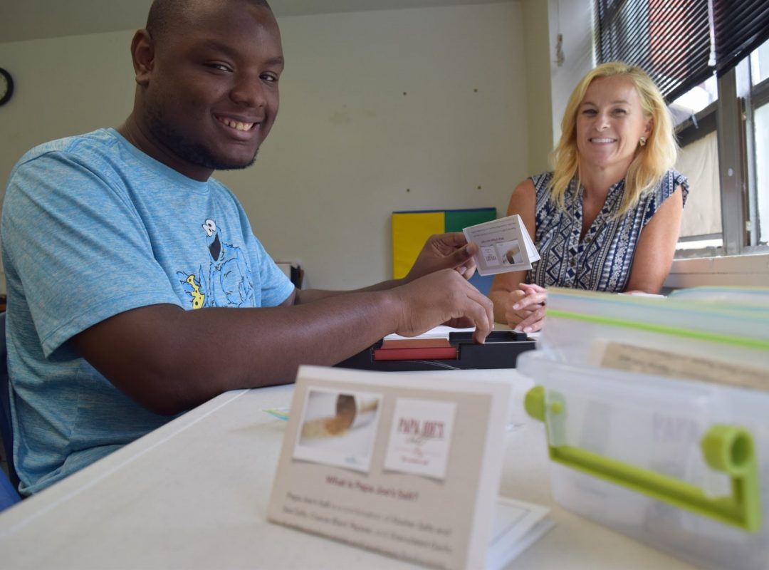 a person served sits in a blue cookie monster shirt holding a card out to a female paraprofessional in a blue top, smiling