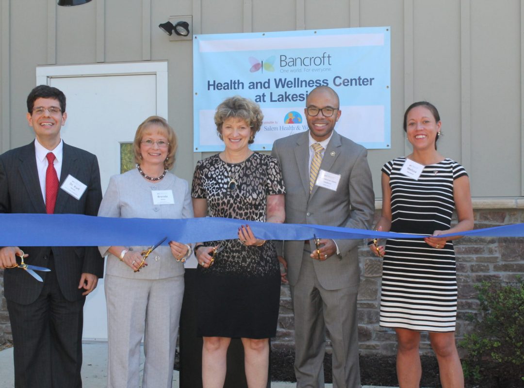 five Bancroft board members stand holding a blue ribbon and large scissors outside of the new Health and Wellness Center at Lakeside building