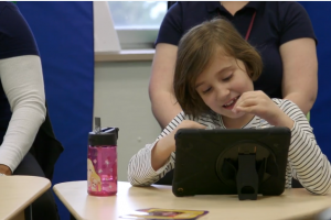 Sophia, a young girl with brown hair and a cream top, sits at a desk with paraprofessionals on a computer