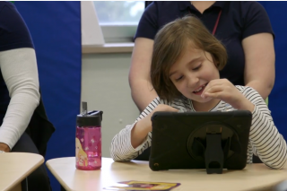 Sophia, a young girl with brown hair and a cream top, sits at a desk with paraprofessionals on a computer