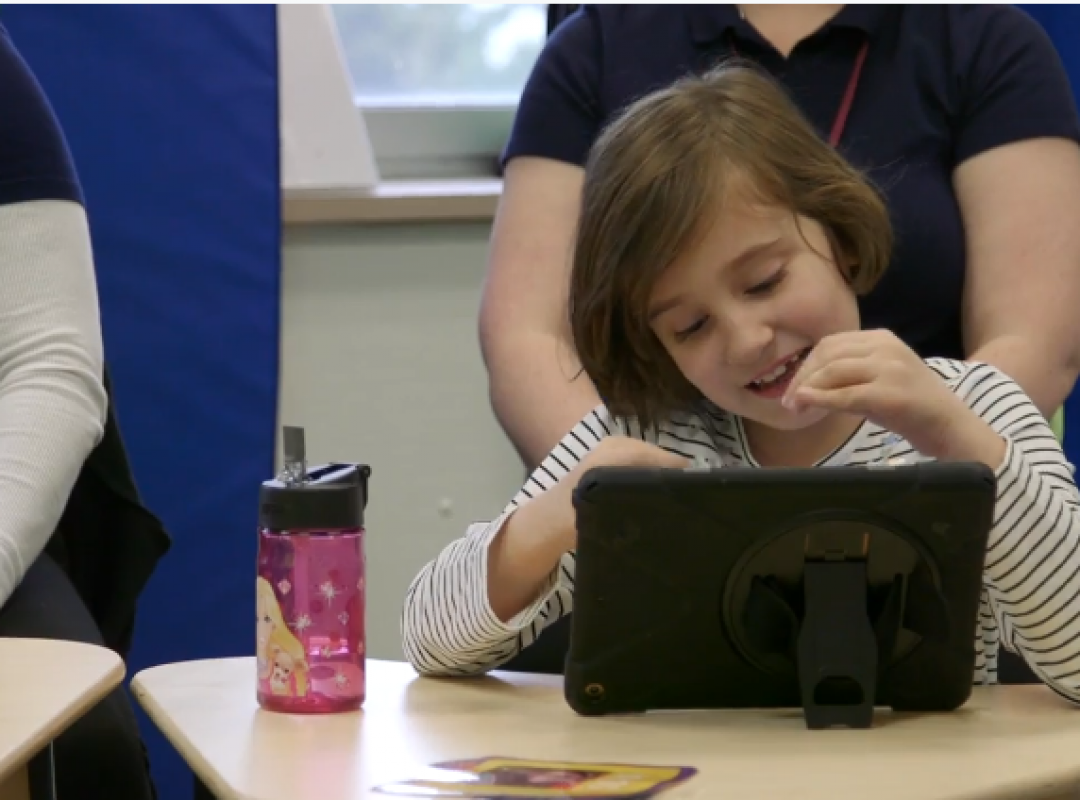 Sophia, a young girl with brown hair and a cream top, sits at a desk with paraprofessionals on a computer