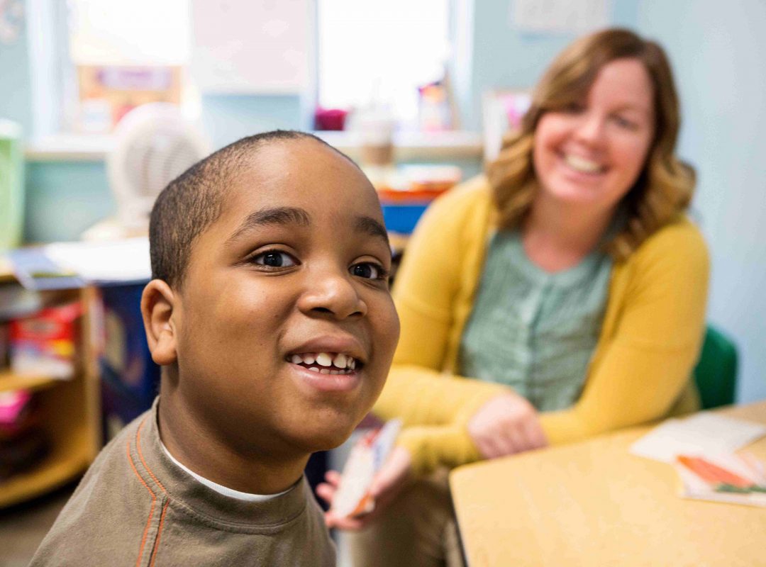 a young boy in a brown shirt sits at a table and smiles while a female paraprofessional in a yellow cardigan watches him, smiling